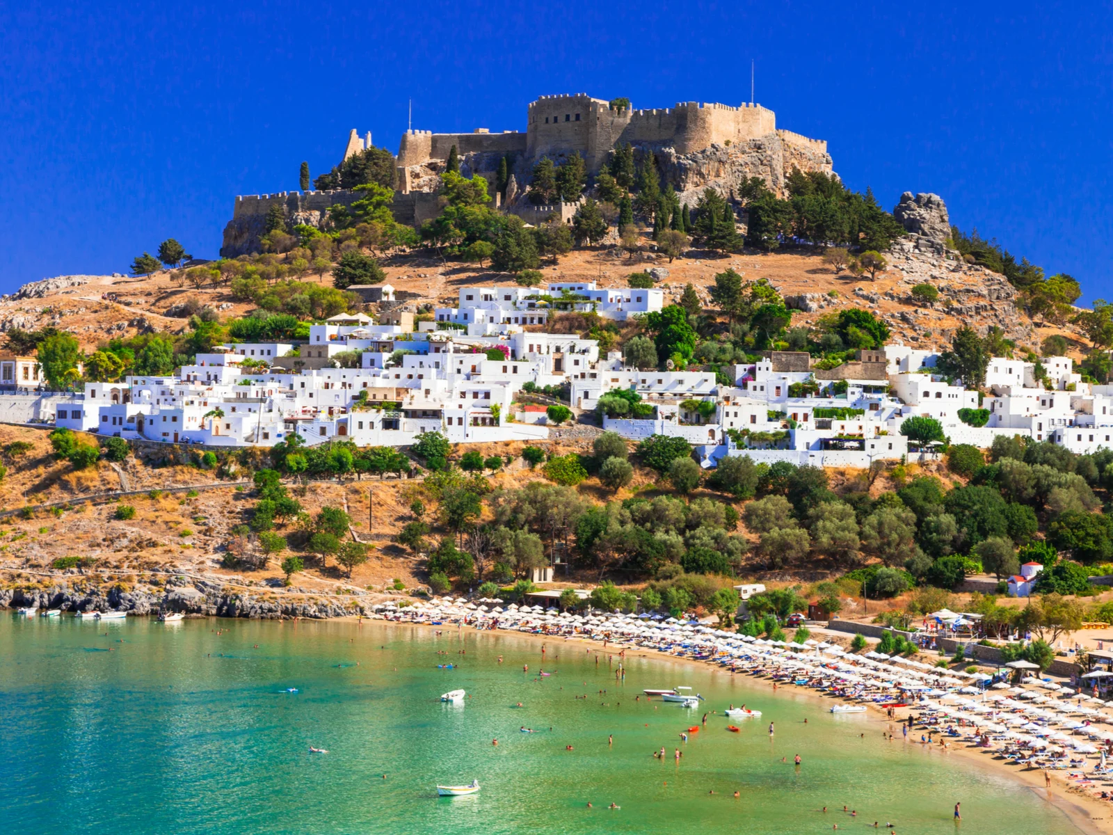 And old castle sitting on a mountain peak and white residential and commercial buildings at one of the best islands in Greece to visit, Rhodes Island with its busy beach during summer season