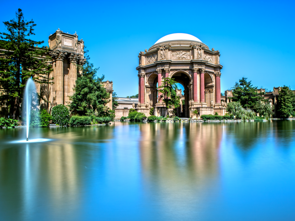 Palace of Fine Arts reflected on the still reflection pool with an outlet of spouting water on its left, pictured with a clear blue sky as a piece on best places to visit in San Francisco