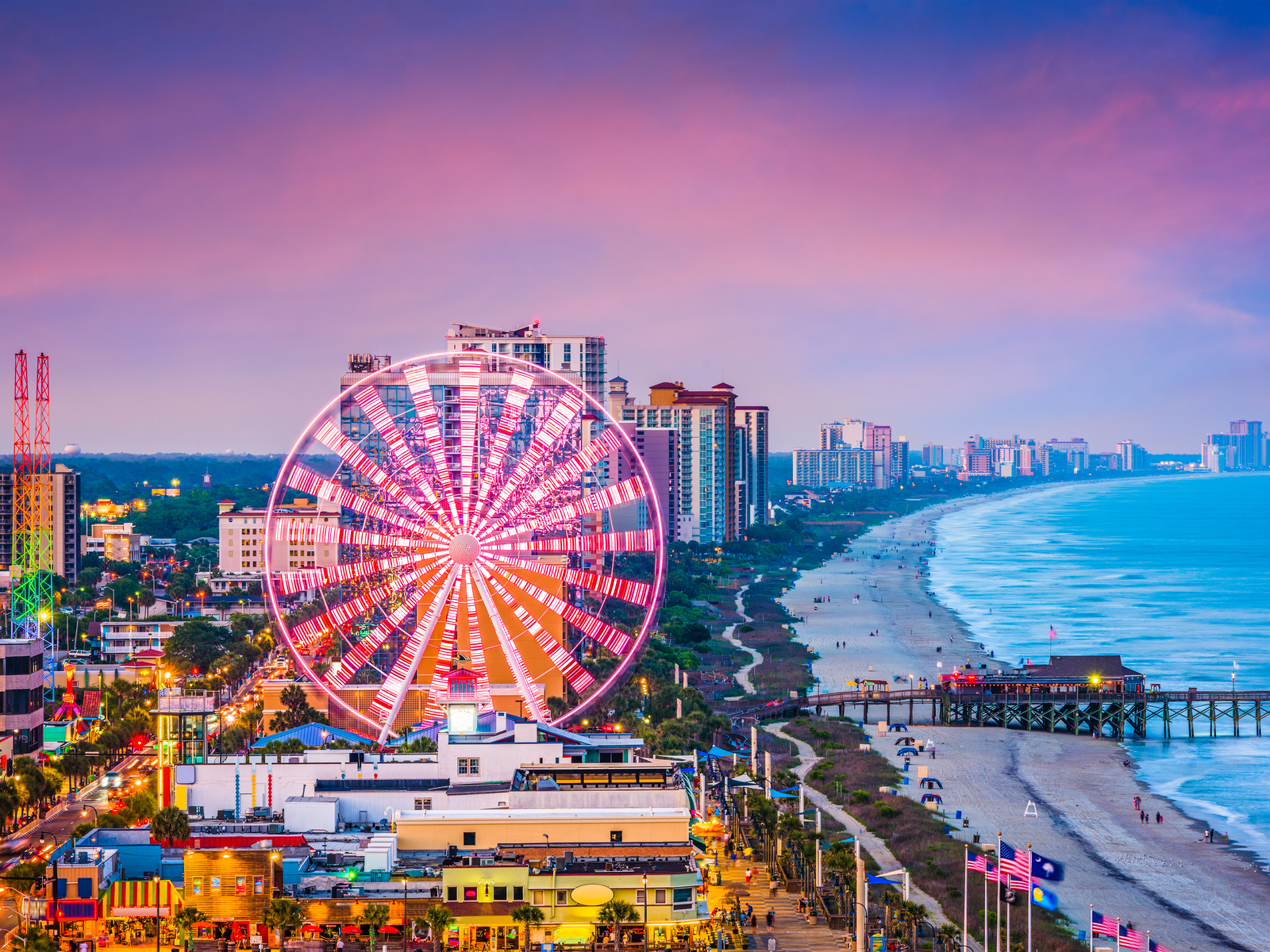 Scenic aerial view on Myrtle Beach's tourists strolling on the shore beside towering hotel buildings and iconic spinning Skywheel, one of the best South Carolina attractions on a late sunset
