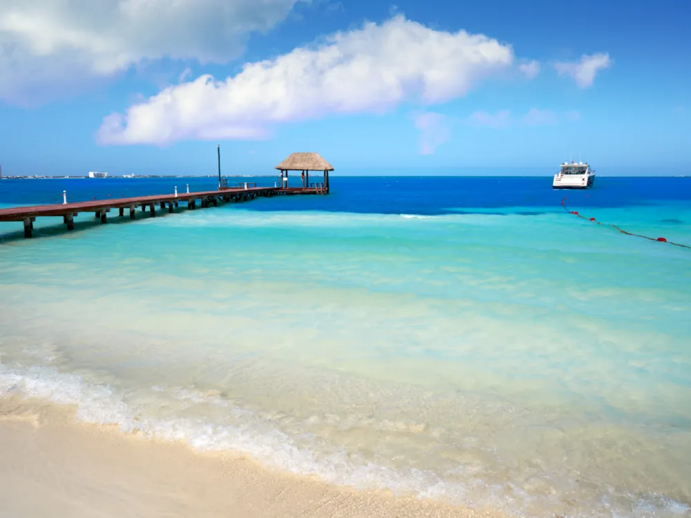 A hut at the end of a boardwalk and a ship anchored at a deep portion of Playa Langosta, pictured as a piece on the best beaches in Cancun