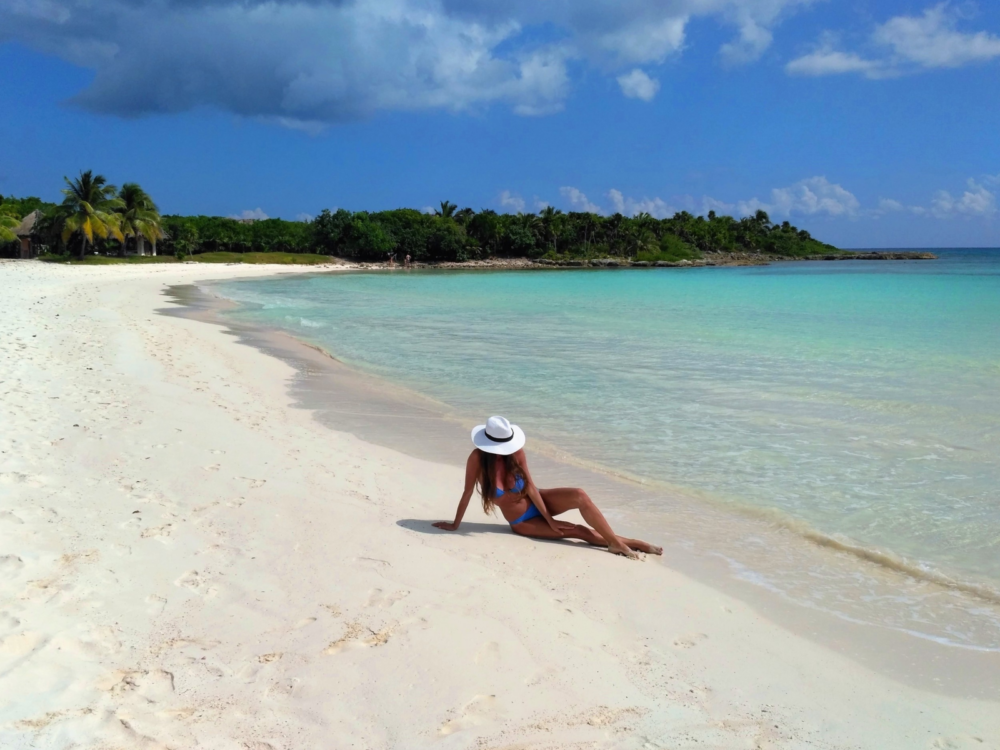 A tan young woman on hear swimwear and white sun hat posing on fine sand during a bright summer day at Xpu-ha Beach, one of the best beaches in Cancun