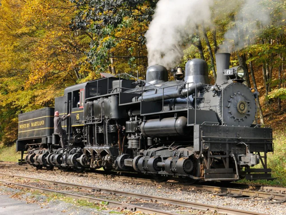 A classic steam engine train blowing off steam while running, one of the best attractions in West Virginia, on rail beside trees on Autumn