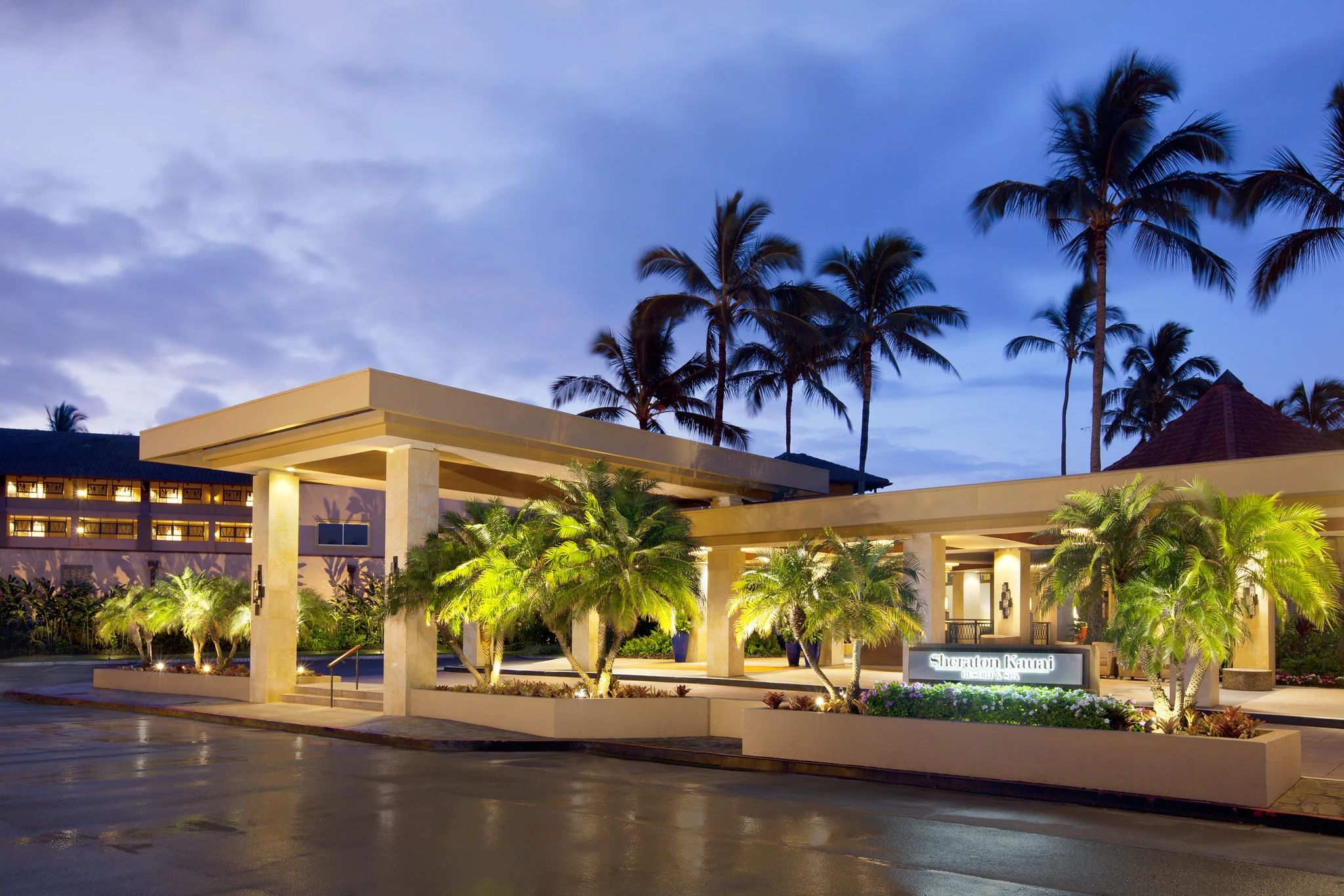 The entrance on Sheraton Kauai Coconut Beach Resort at dusk, a piece on the best hotels in Kauai, with lit sign beside decorative plants