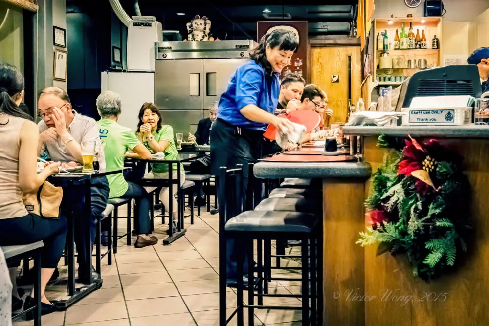 A female service crew wearing her blue uniform smiling while cleaning the counter table, and several tourist diners enjoying the Sushi at Furusato Sushi, a Japanese restaurant in Hawaii and one of the best restaurants in Oahu