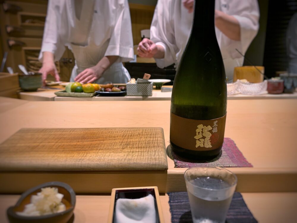 Chefs preparing a Shushi at the counter having all the ingredients, and a traditional wooden board plate sits beside a Kokuryu Ryu Daiginjo "Gold Dragon" Sake at Sushi Sho, a Japanese restaurant and one of the best restaurants in Oahu, Hawaii