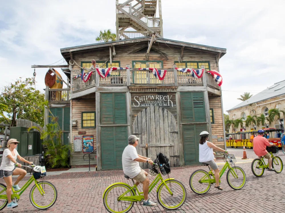 Four people riding green bicycles passing by Shipwreck Treasure Museum, one of the best things to do in Key West