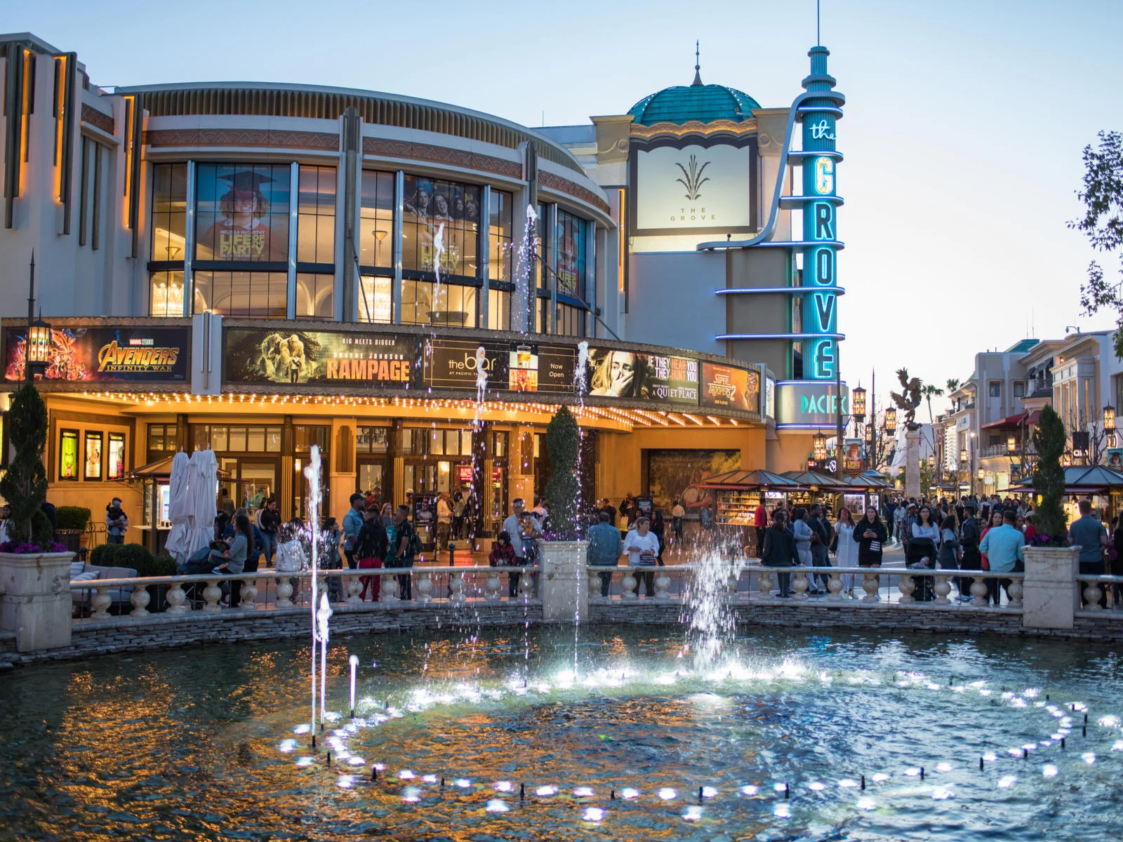 A number of people walking by the a large fountain in front of the lively The Grove Mall in Los Angeles with movie posters above the entrance, one of the best malls in America