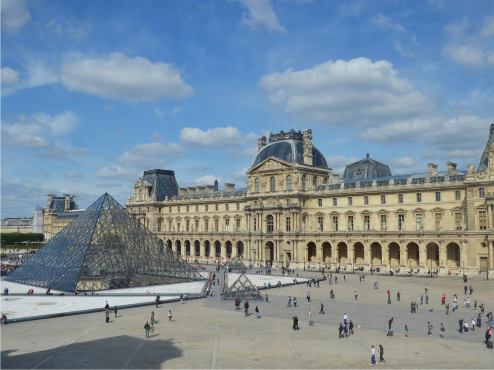 The Louvre pictured on a slightly sunny day with a bunch of people in the front gathering area