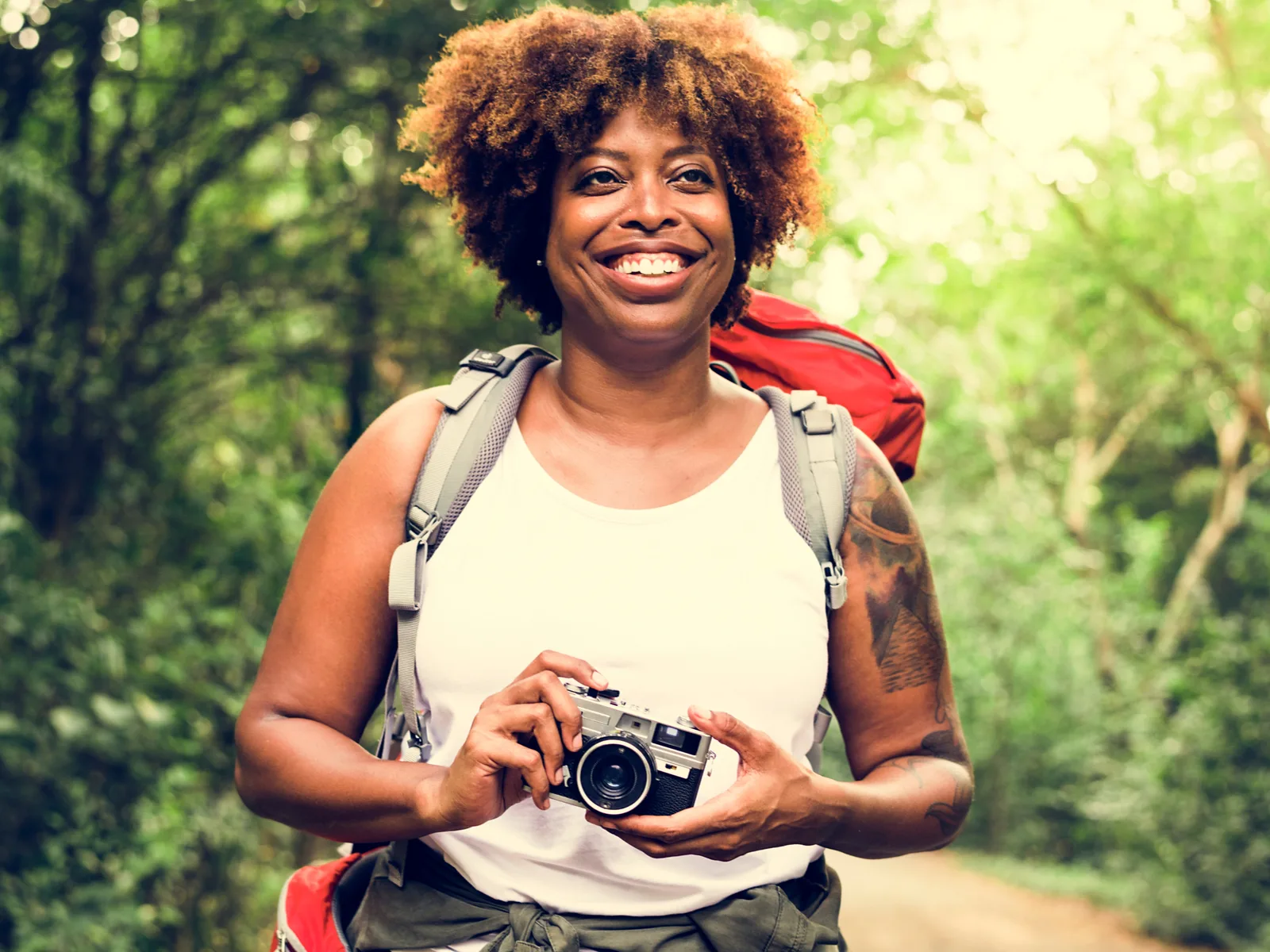 Woman with natural hair smiling as she walks through the woods on a hiking path wearing one of the best hiking camera backpacks that's red in color