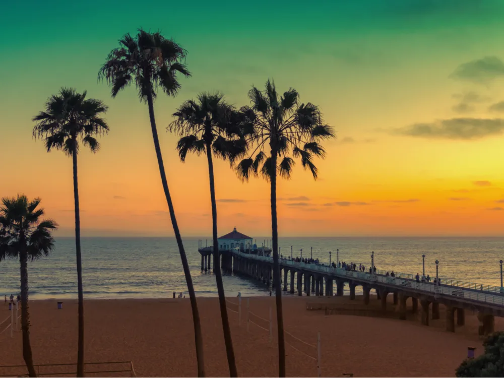 A sweet sunset over people walking along the long Venice Beach Boardwalk with a silhouette of palm trees, a piece on the best things to do in California