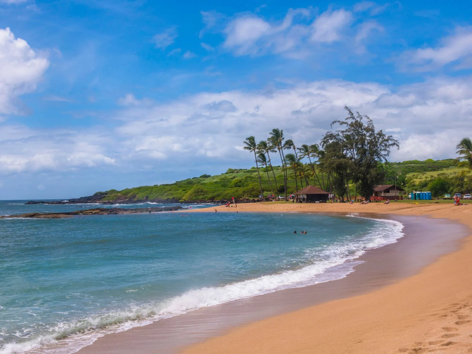 Three tourists swimming in the turquoise waters of Salt Pond Beach, considered one of the best beaches in Kauai, onshore are two beach houses beside palm trees