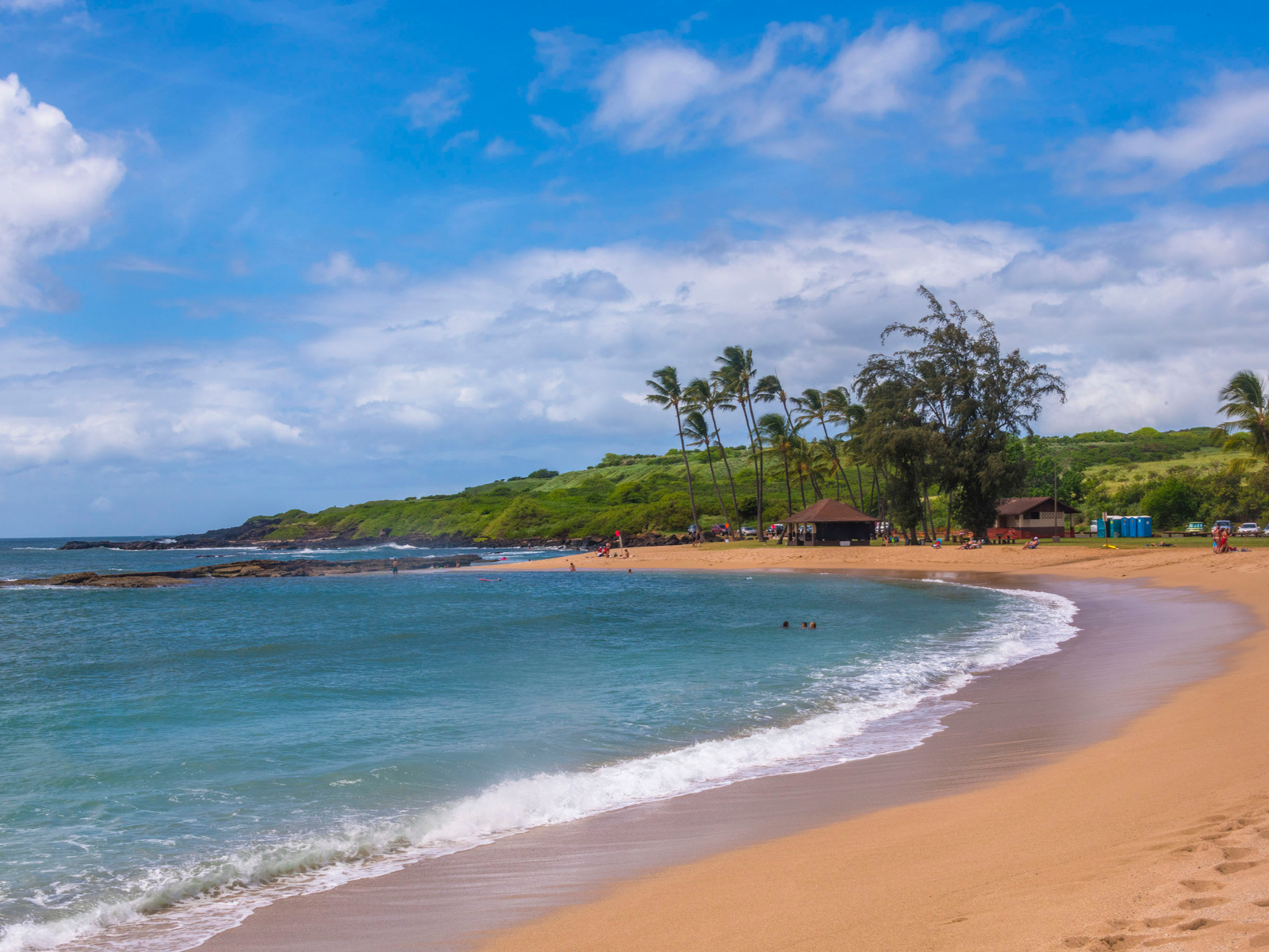 Visitors enjoying the calm waves at Salt Pond Beach with two beach houses near a group of palm trees, one of the best snorkeling spots in Kauai