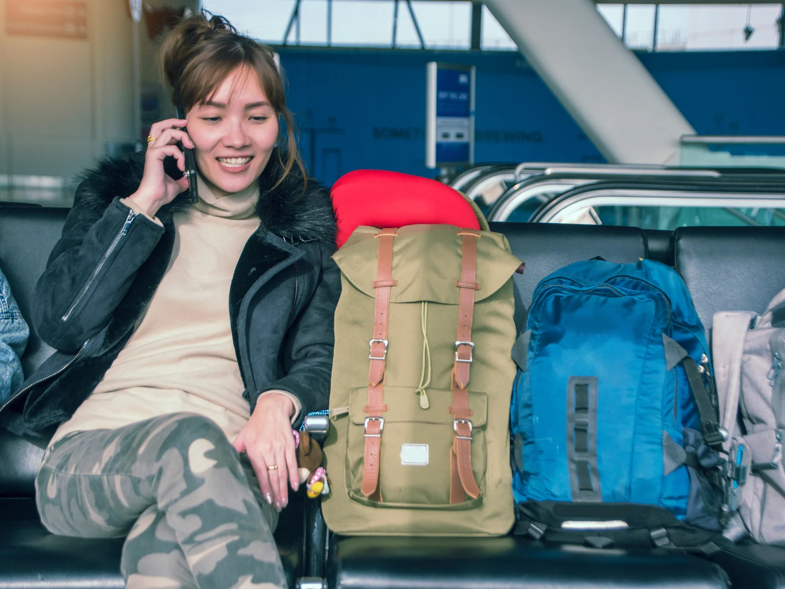 Young Asian woman sitting next to her carry-on luggage while talking on the phone in camo pants