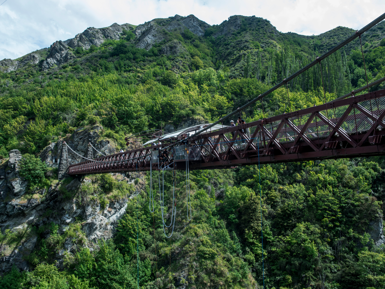 A man preparing to bungee jump together with some friends on Kawarau Suspension Bridge, a green mountain in background, that is one of the most famous Lord of the Rings filming locations