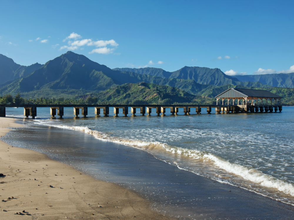 The empty pier at Hanalei Bay, one of the best snorkeling spots in Kauai, with its brown sand and a beautiful mountain landscape in the background
