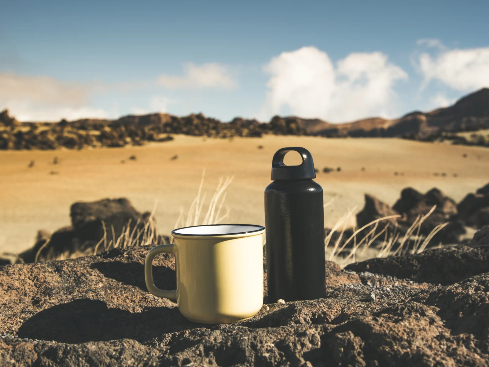Close up of one of the best insulated water bottles next to a ceramic mug on the rocks at a remote camping site