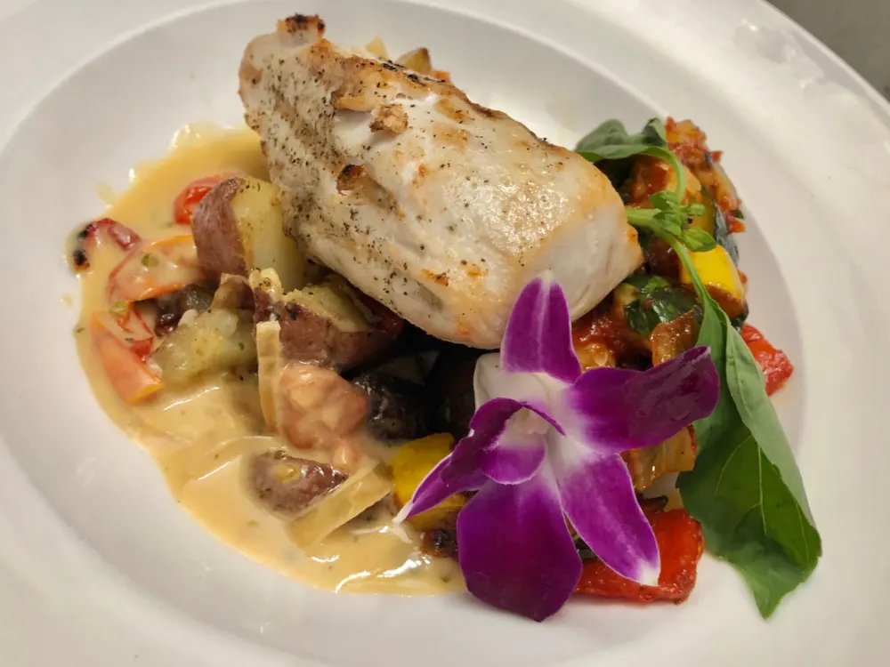 A chunk of freshly caught Wahoo, grilled and served with cream sauce, on a white plate together with a flower at Ono Seafood, a Hawaiian seafood restaurant and one of the best restaurants in Oahu
