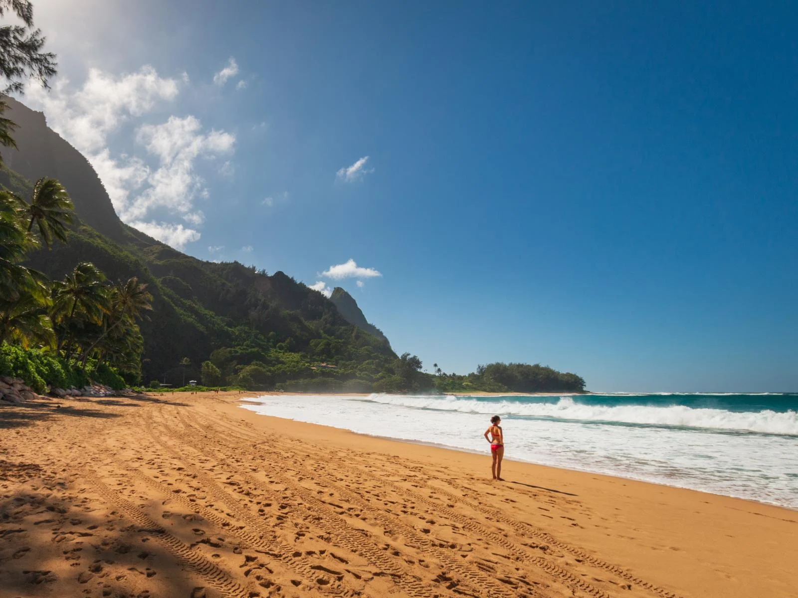 A woman in her summer swimwear standing on the golden sand while putting both hands on her hips facing the wavy sea at Makua Tunnels Beach, a piece on one of the best beaches in Kauai