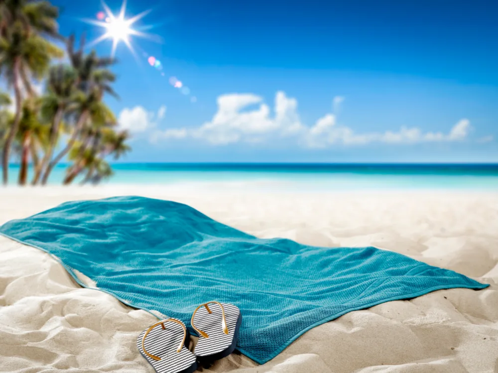 Image of the best travel towel (green in color) lying on a beach next to palm trees and flip flops