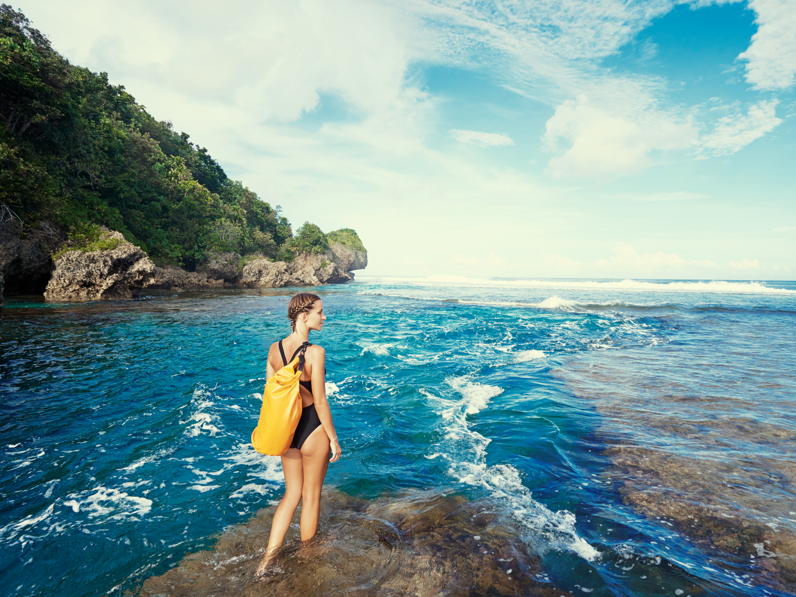Thin woman in a one-piece swimsuit wearing one of the best waterproof backpacks and standing on coral by the ocean