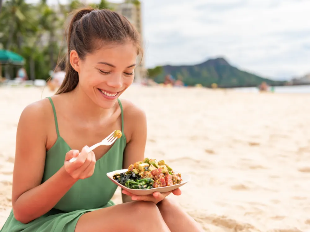 Woman eating Poke on the beach for a piece on the best restaurants in Oahu