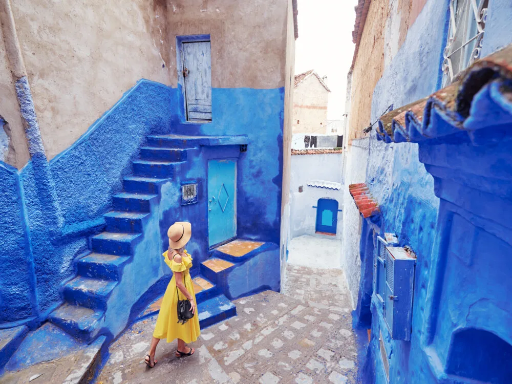 Thin woman in a yellow dress walking through the colorful blue and white walls of Fez during the best time to go to Morocco
