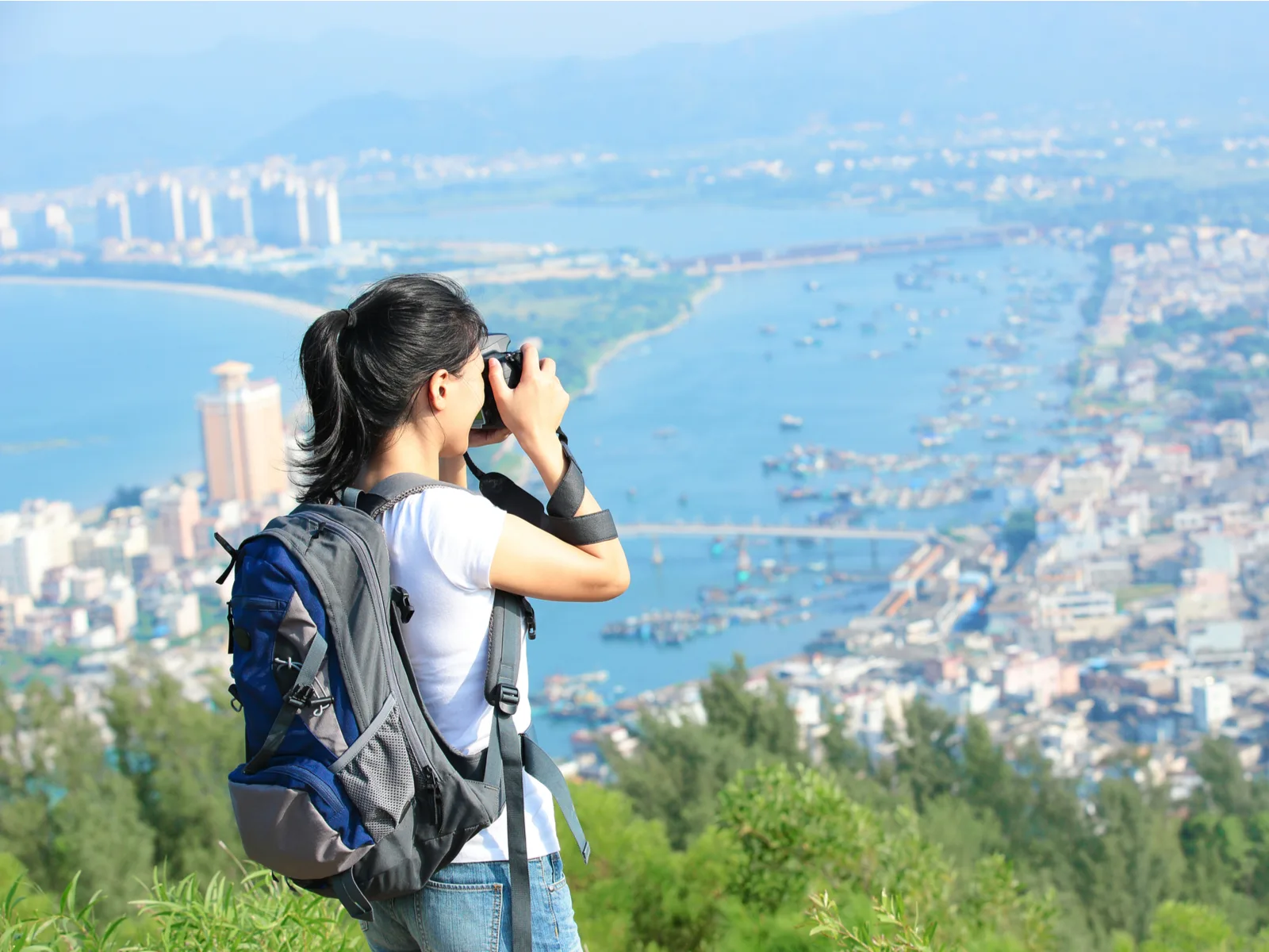 Woman using one of the best hiking camera backpacks while taking a photo of a city below the hill on which she stands