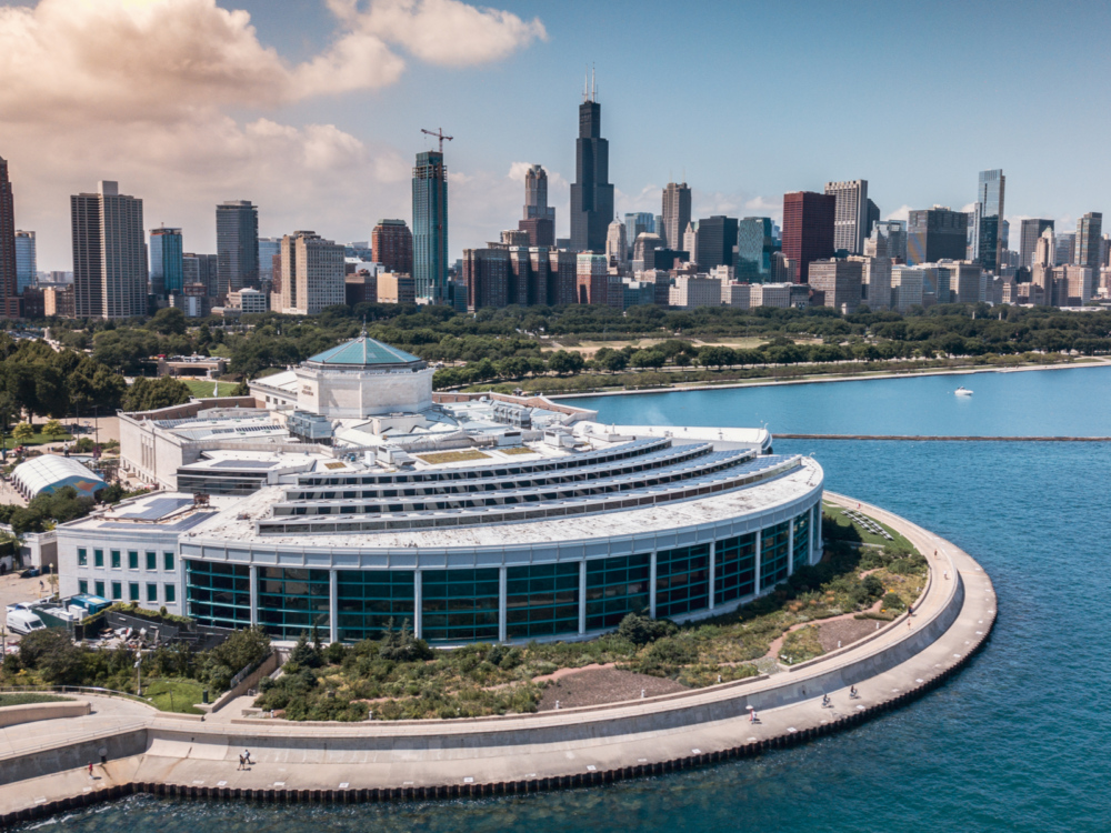 Aerial view of the rounded architecture on Shedd Aquarium, one of the best aquariums in the US, with the Chicago skyline in the background and a few people walking along the seaside pathway