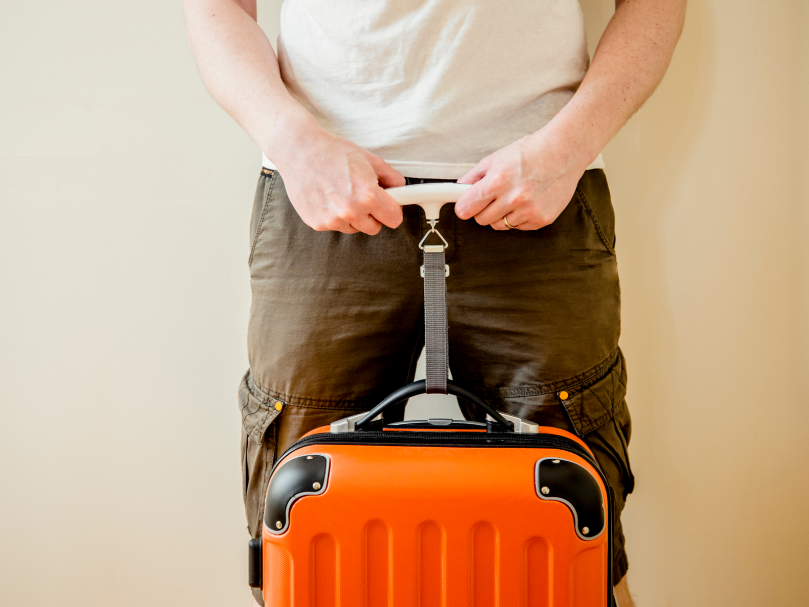Person holding an orange piece of luggage that's hanging from one of the best luggage scales in a beige room while wearing hiking pants