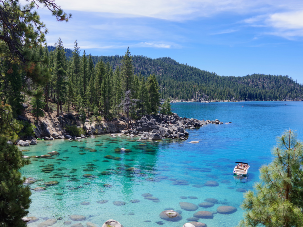 During the best time to visit Lake Tahoe, the Nevada side pictured with a pontoon boat floating above some rocks with pines all around