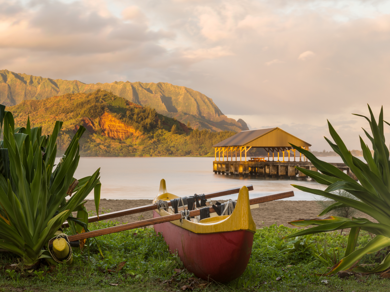 A red canoe with an outrigger on the shore and the Hanalei pier on an early morning at Hanalei Bay Beach, considered one of the best beaches in Kauai