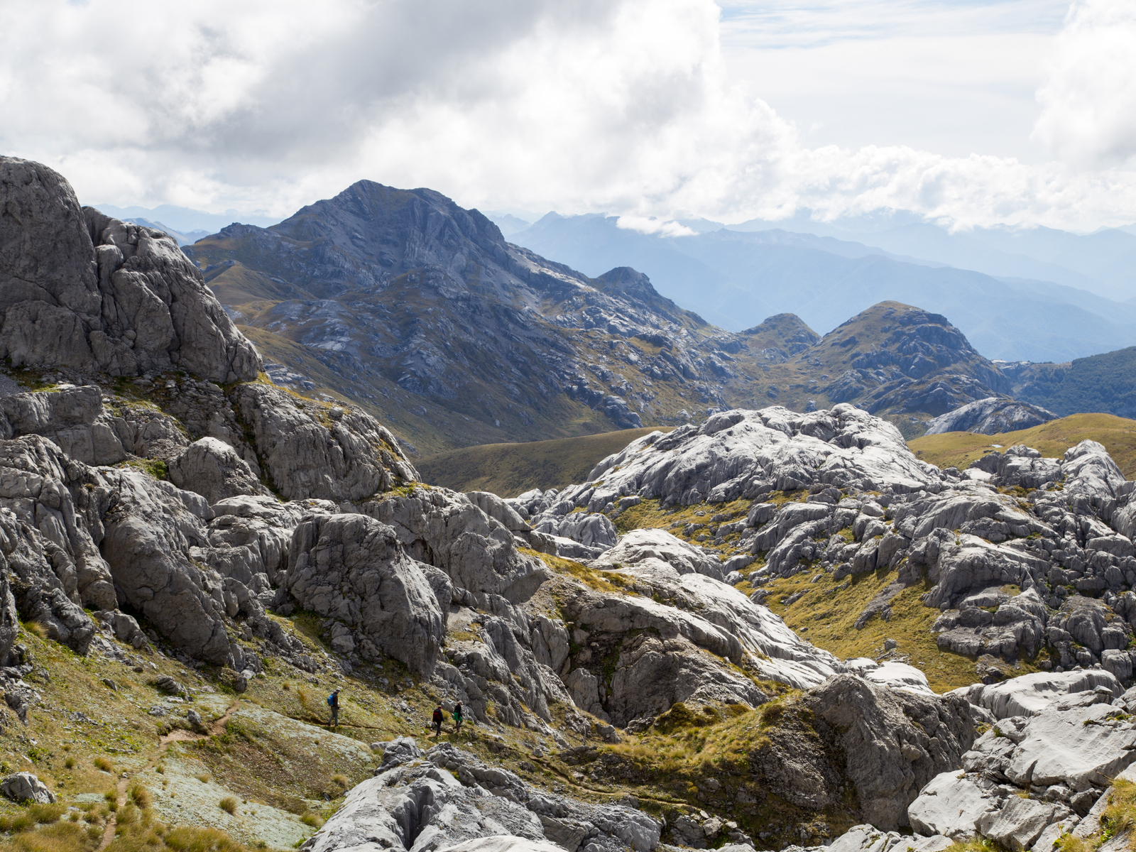 Aerial photo of three people hiking down a rocky trail in Mount Owen during a cloud day and the skyline of Kahurangi National Park, one of the Lord of the Rings filming locations you can visit