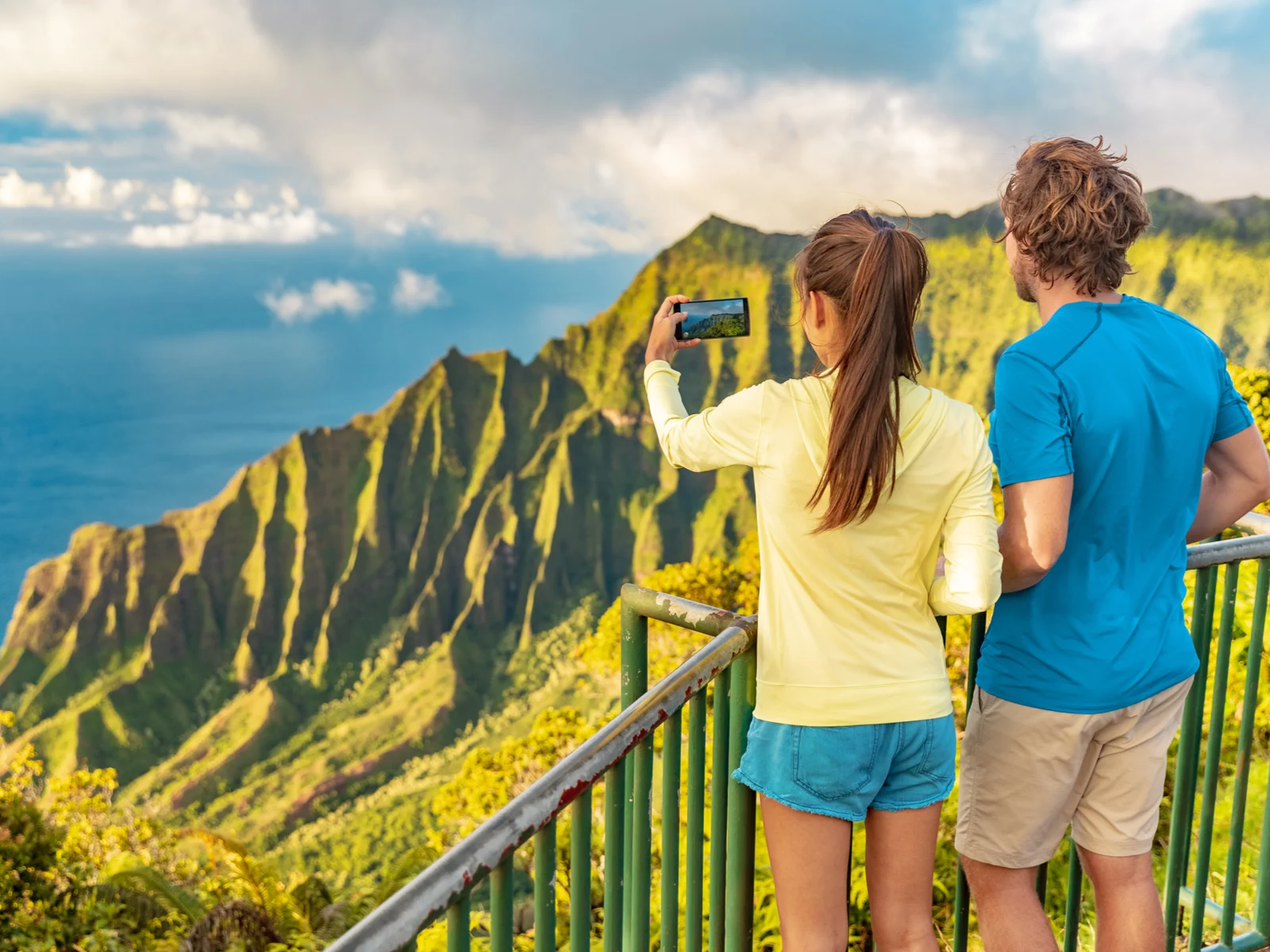 A couple taking a picture of the beautiful landscape of Na Pali Coast mountain range, one of the best things to do in Kauai