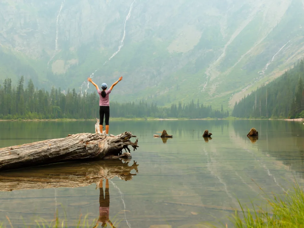 Slender Asian woman holding her arms up over a lake in the mountains during the least busy time to visit Montana featuring fog in the valley