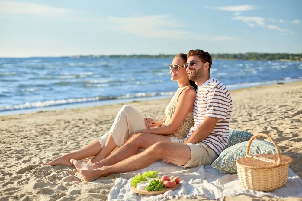 Couple sitting on one of the best picnic blankets on a beach looking out over the water under a clear sky