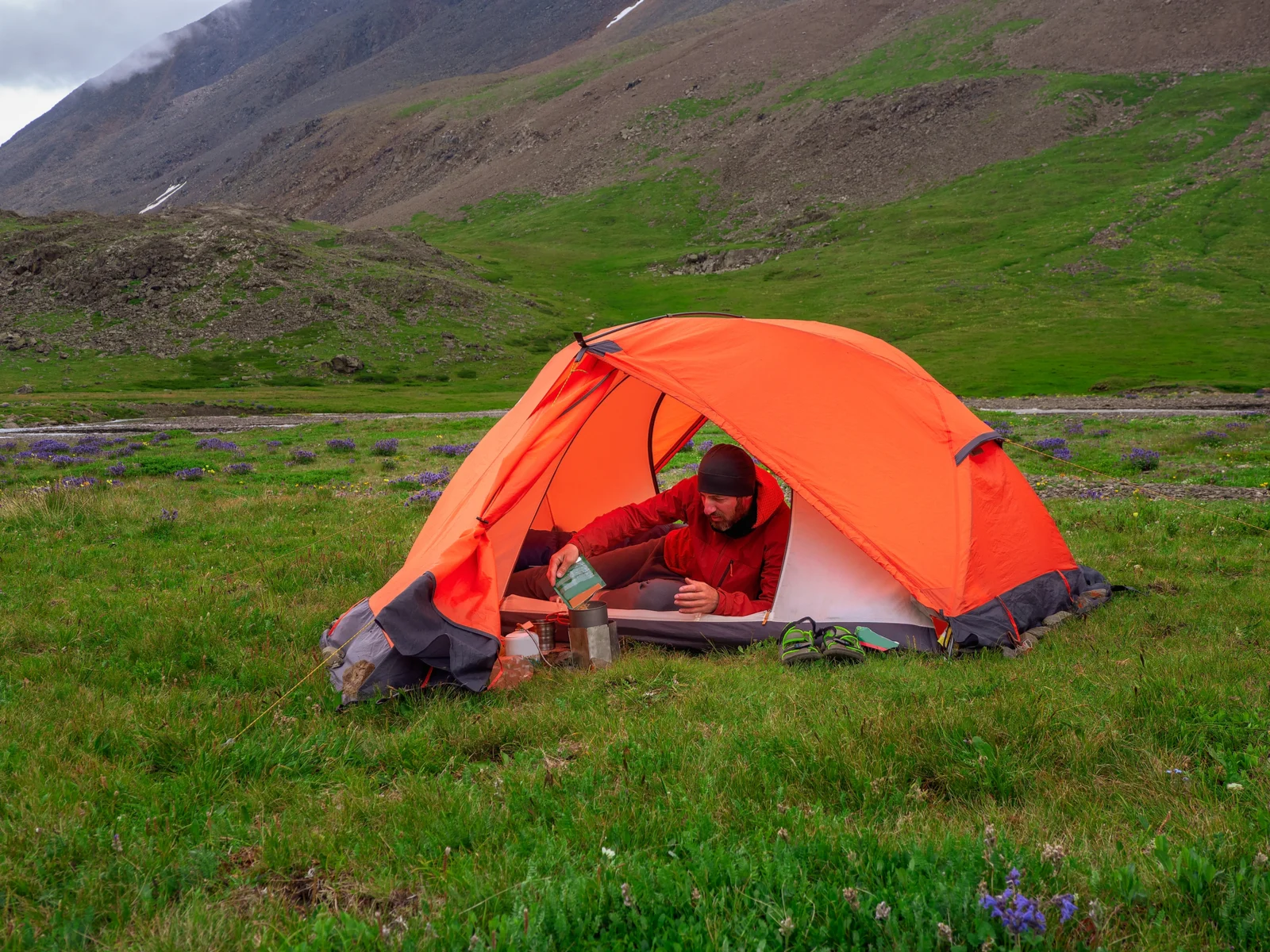 Guy in a hat and and orange tent pouring coffee into a thermos on green grass with purple flowers