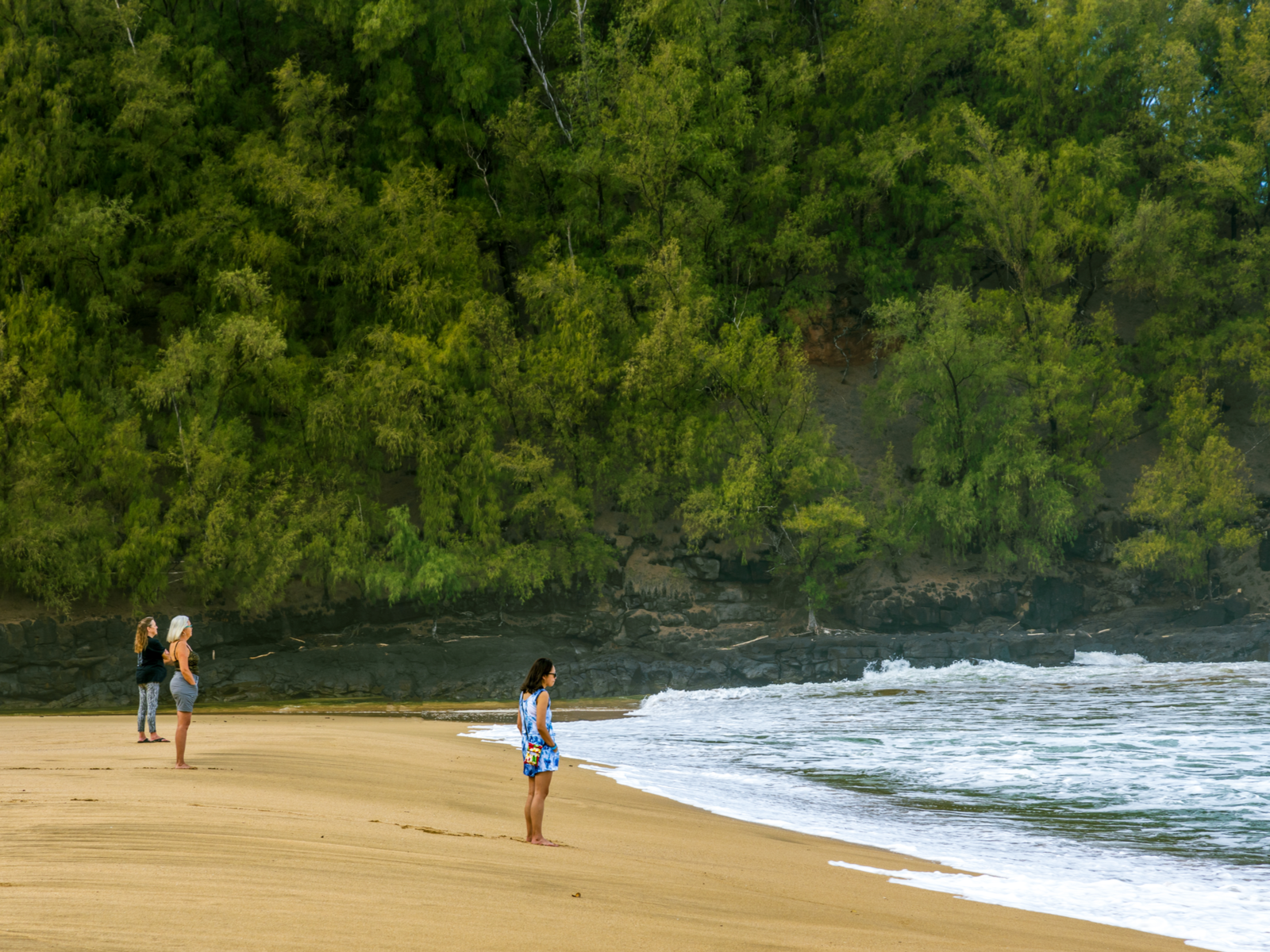 Three women staring into the distant sea of Kalihiwai Beach, one of the best beaches in Kauai, with a slope filled with trees in the background