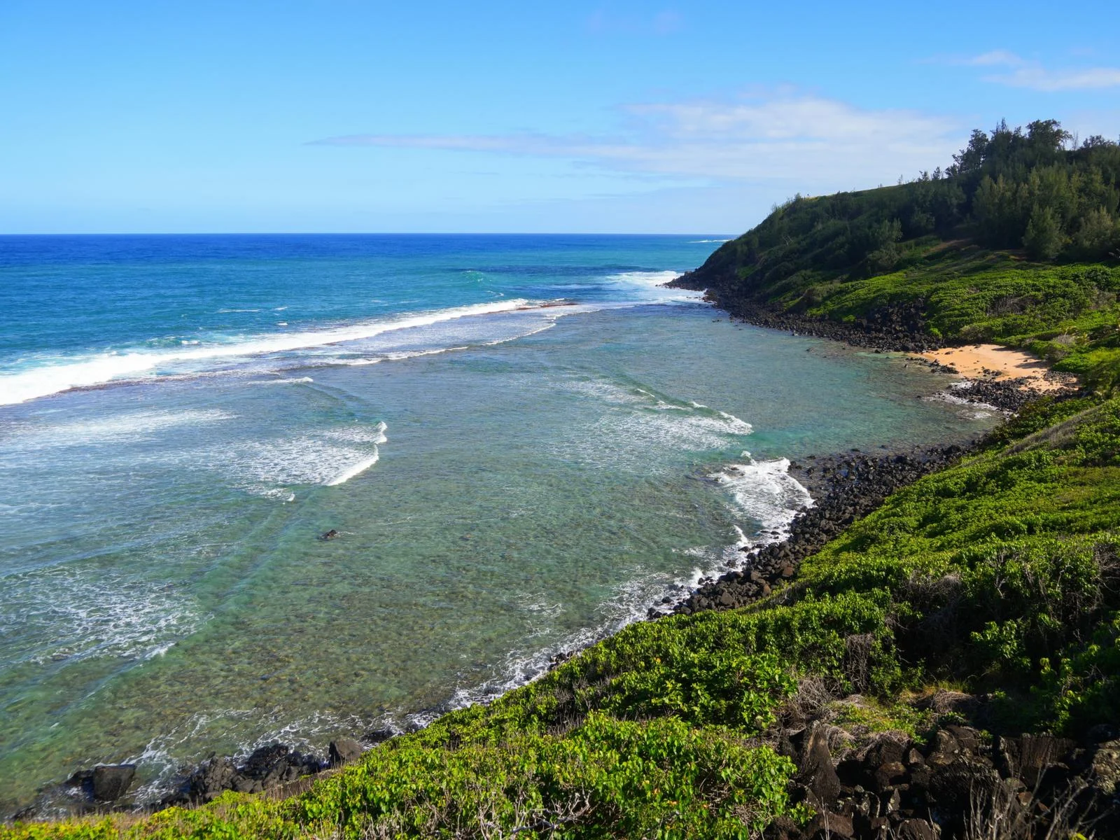 Deep brown rocks at the coast of Larsen’s Beach with a little sand area and bushes growing offshore, considered one of the best snorkeling spots in Kauai