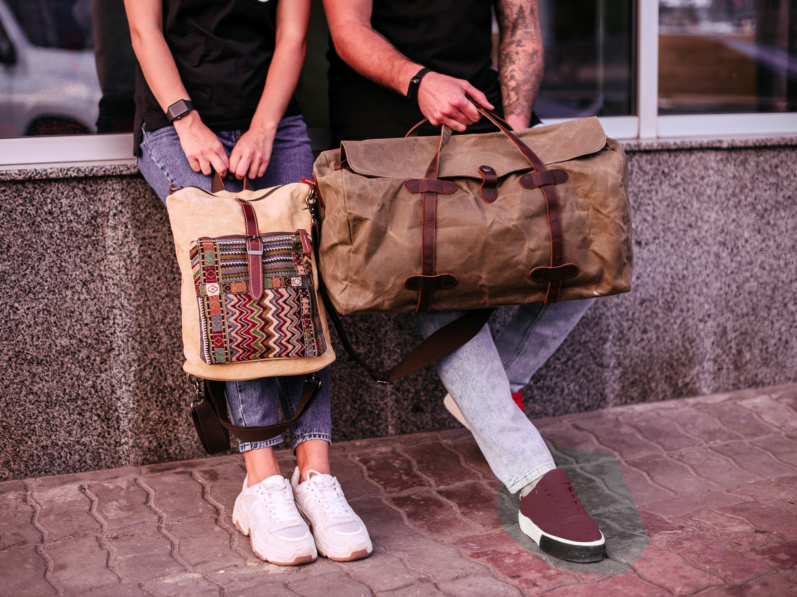 Man and woman holding the best weekender bags while sitting on a marble slab next to a hotel