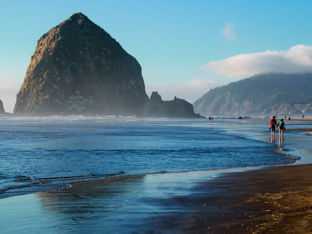 A couple, holding each other's hand, walking along the calm Cannon Beach in Oregon, considered as one of the best beaches in the US, famous for its iconic rock formations