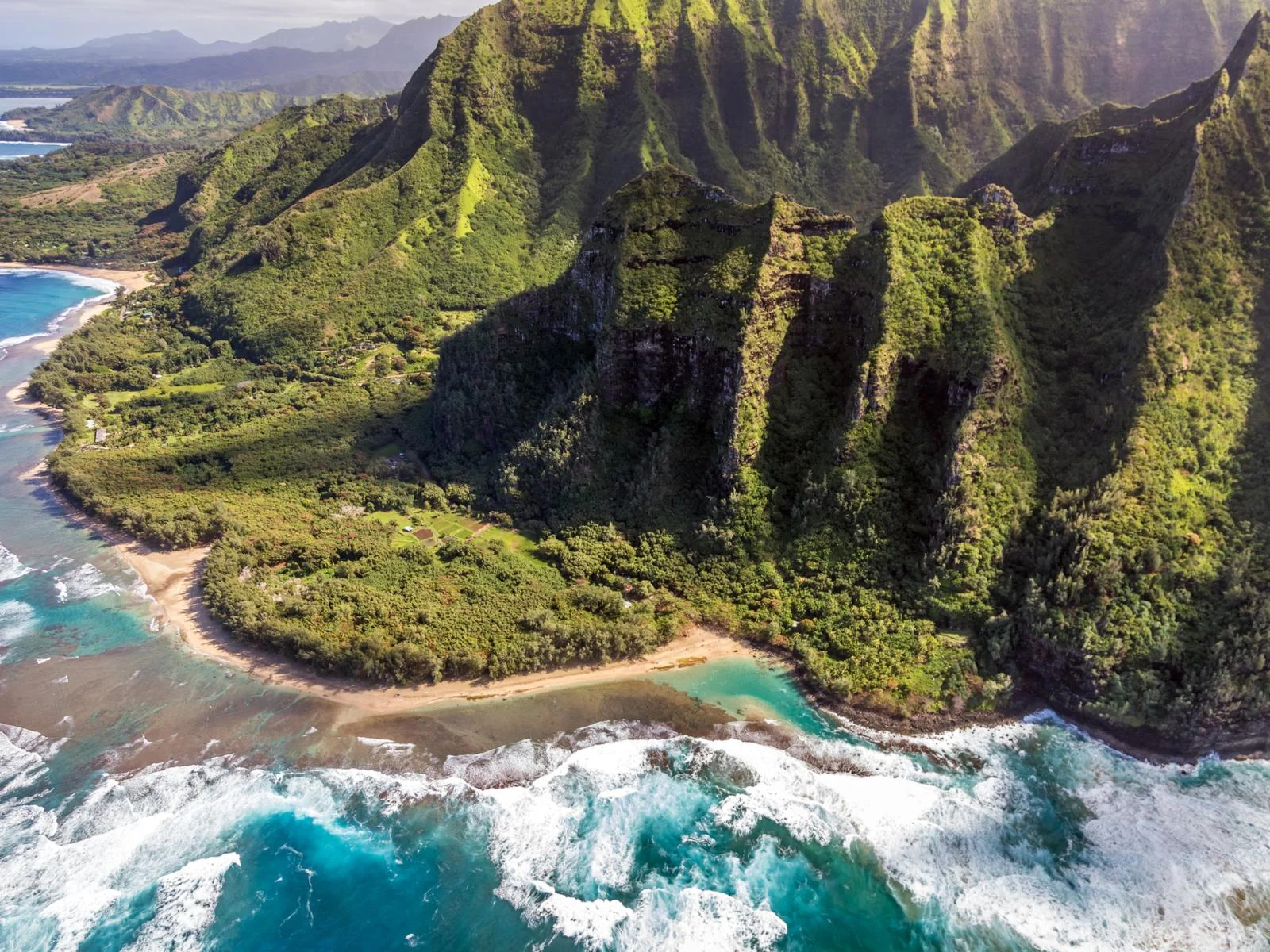 Aerial view of the tall and green mountains of the Na Pali Coast beside the wavy Kee Beach, a piece on the best beaches in Kauai