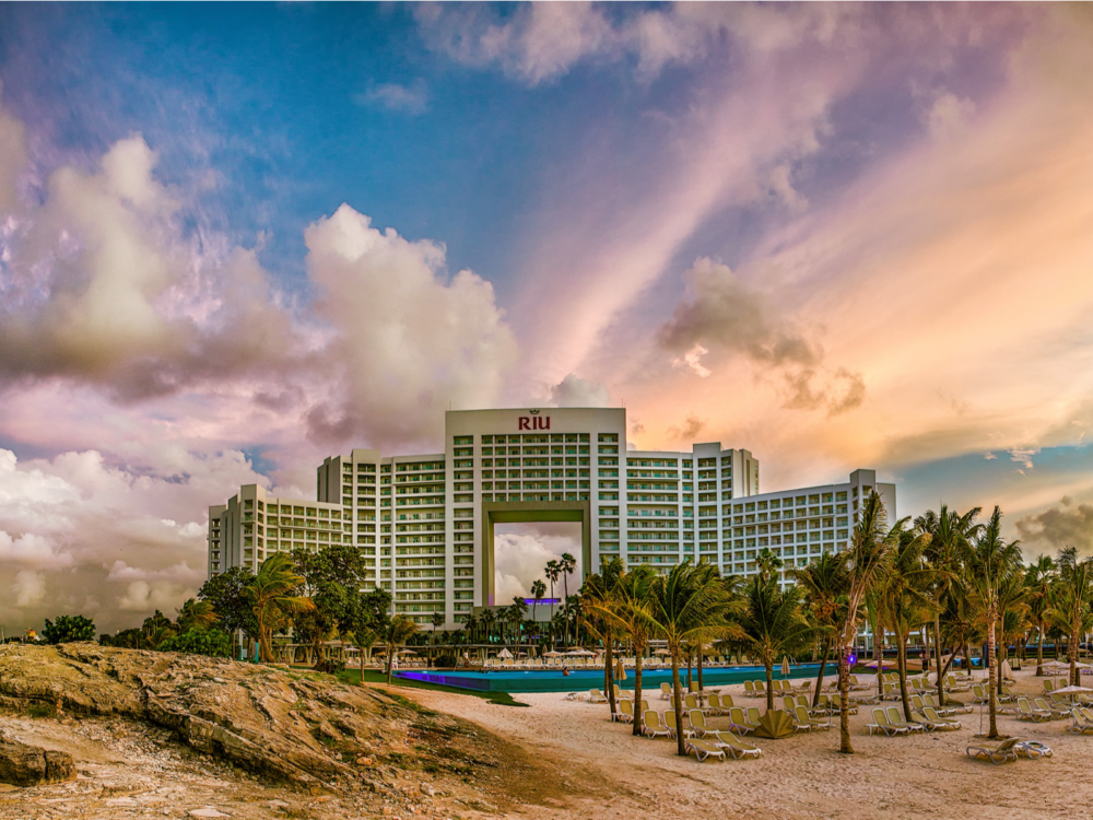 Beautiful sunset over the one of a kind building at Hotel Riu Palace Peninsula, one of the best all-inclusive resorts in Cancun for families, where in front, sunloungers sit underneath palm trees