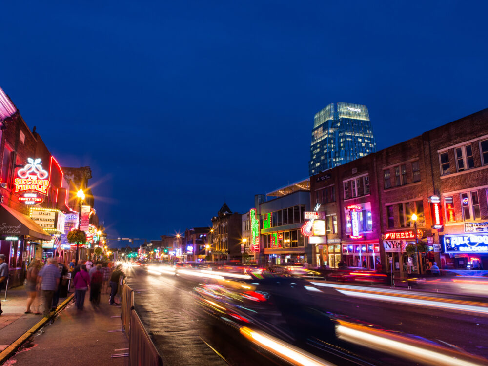 During the best time to visit Nashville, a night shot is taken with a low-frame shot with people walking and the lights of car seen as a blur