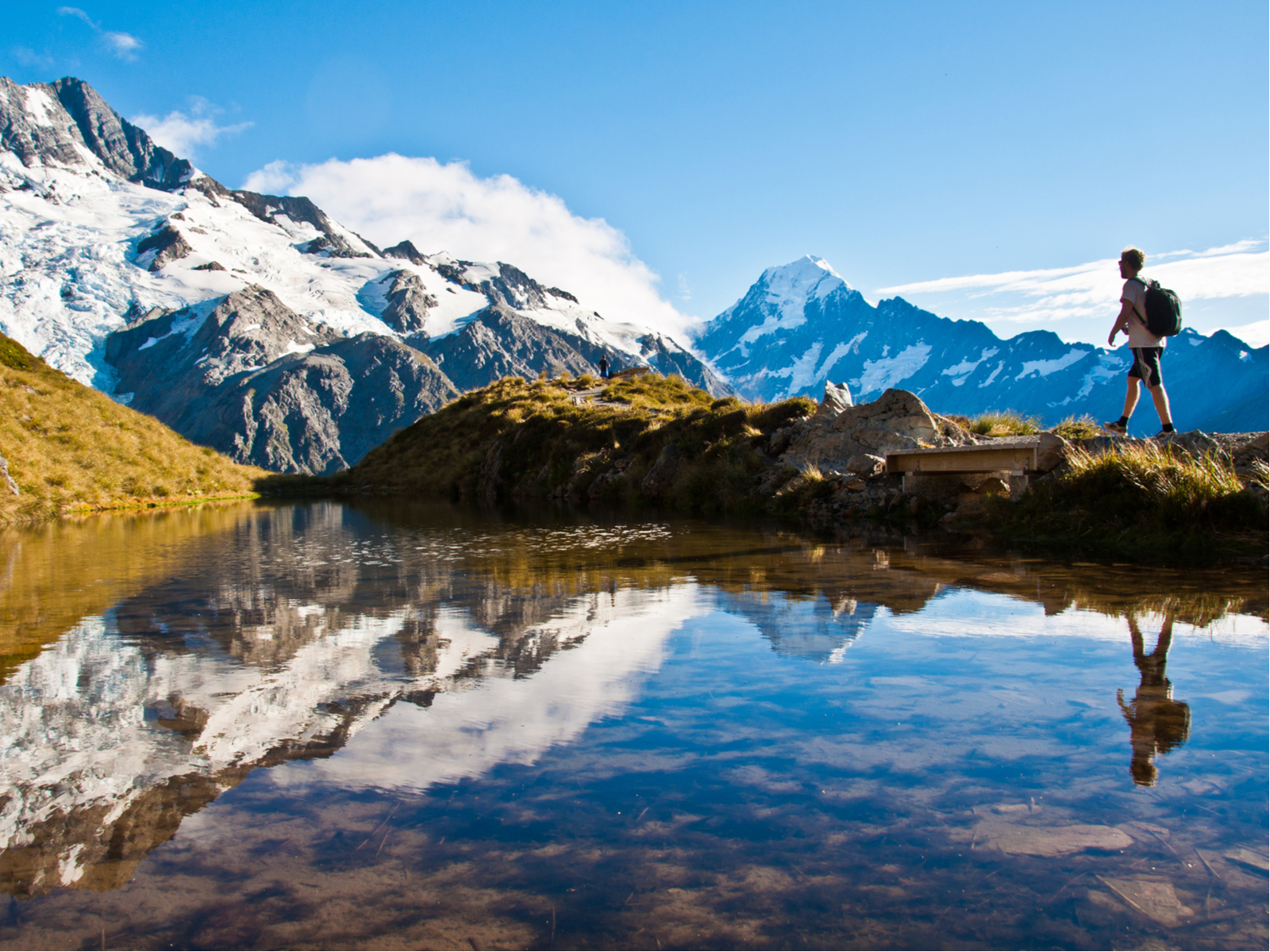 Guy hiking Mount Cook with a day pack and shorts standing in front of a pond with snow-capped mountains in the background