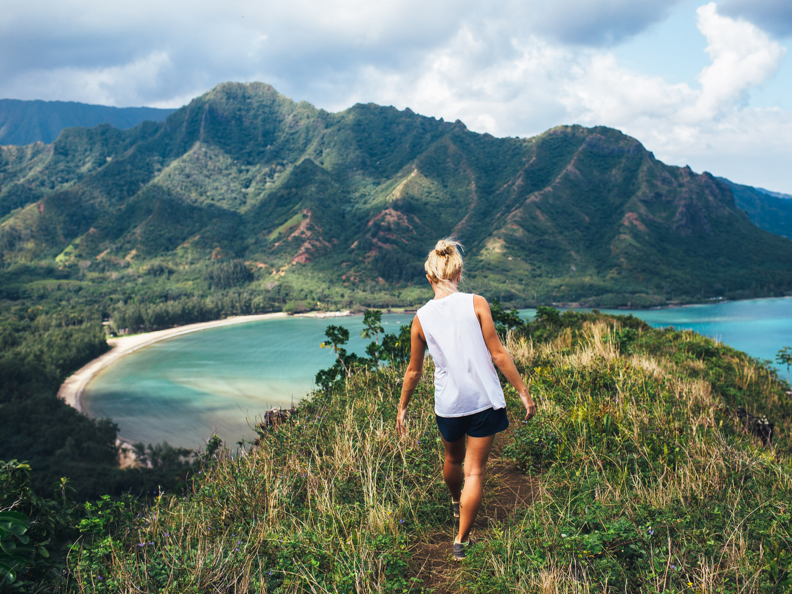 For a post titled Is Hawaii Safe to Visit, a single blonde woman walking on a path overlooking the Na Pali Coast