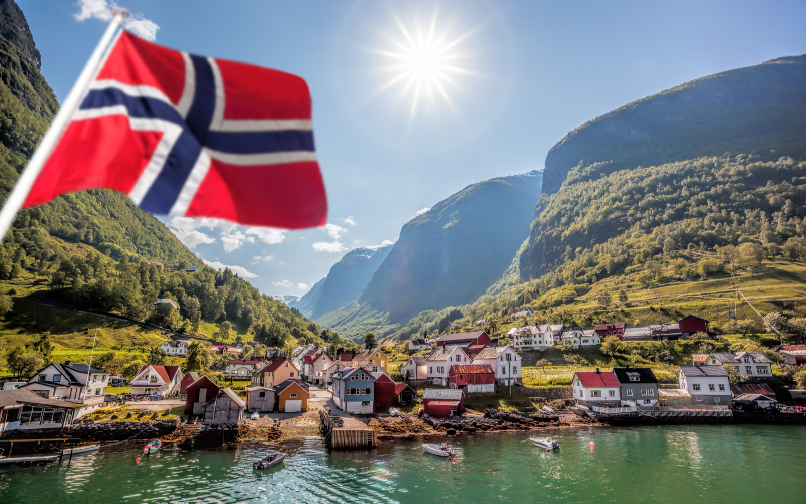 Featured image for a piece on the best time to visit Norway featuring the flag waving in front of a waterside village