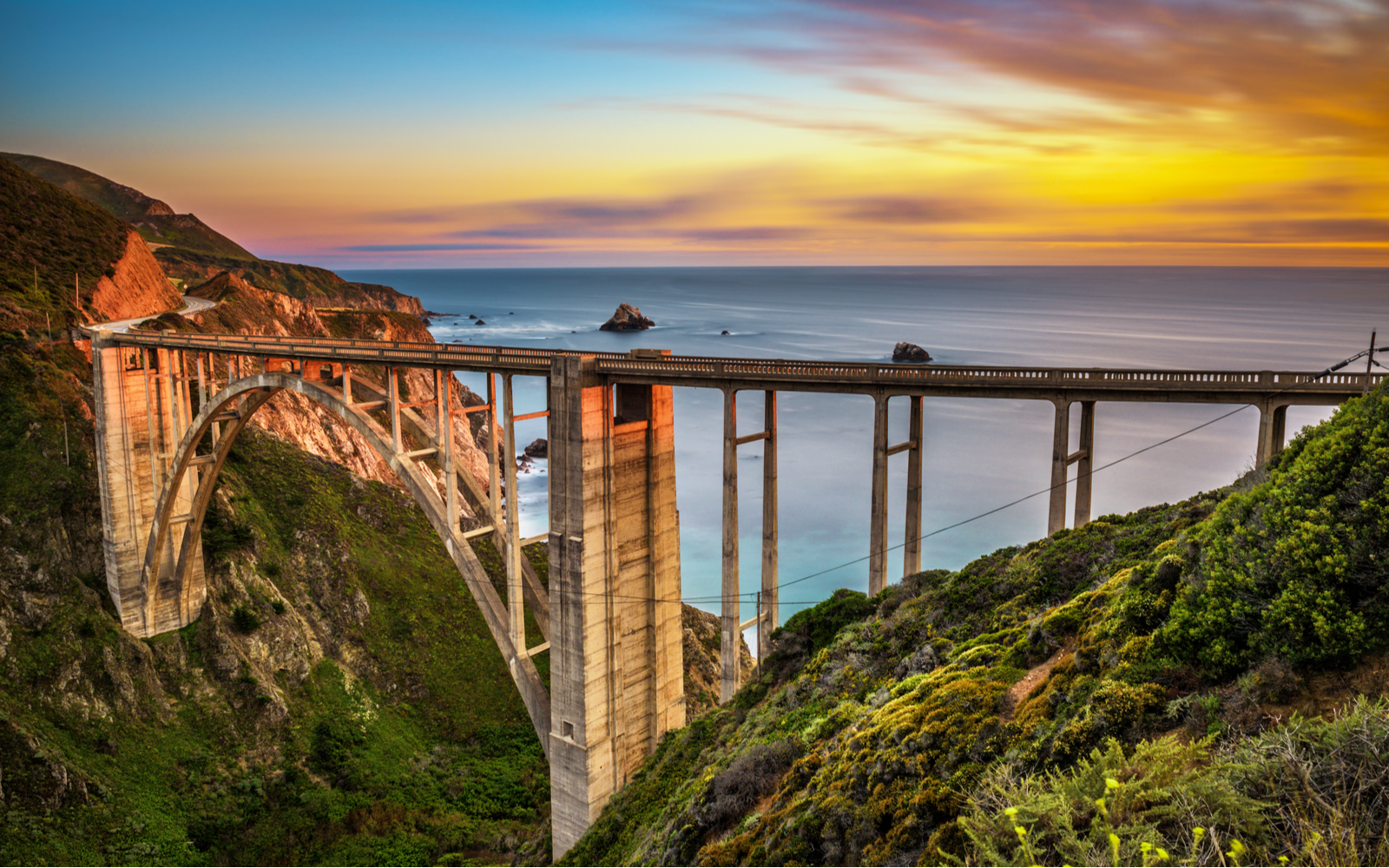 Bixby Bridge pictured above the valley below the sunset over the ocean for a piece on the best things to do in California
