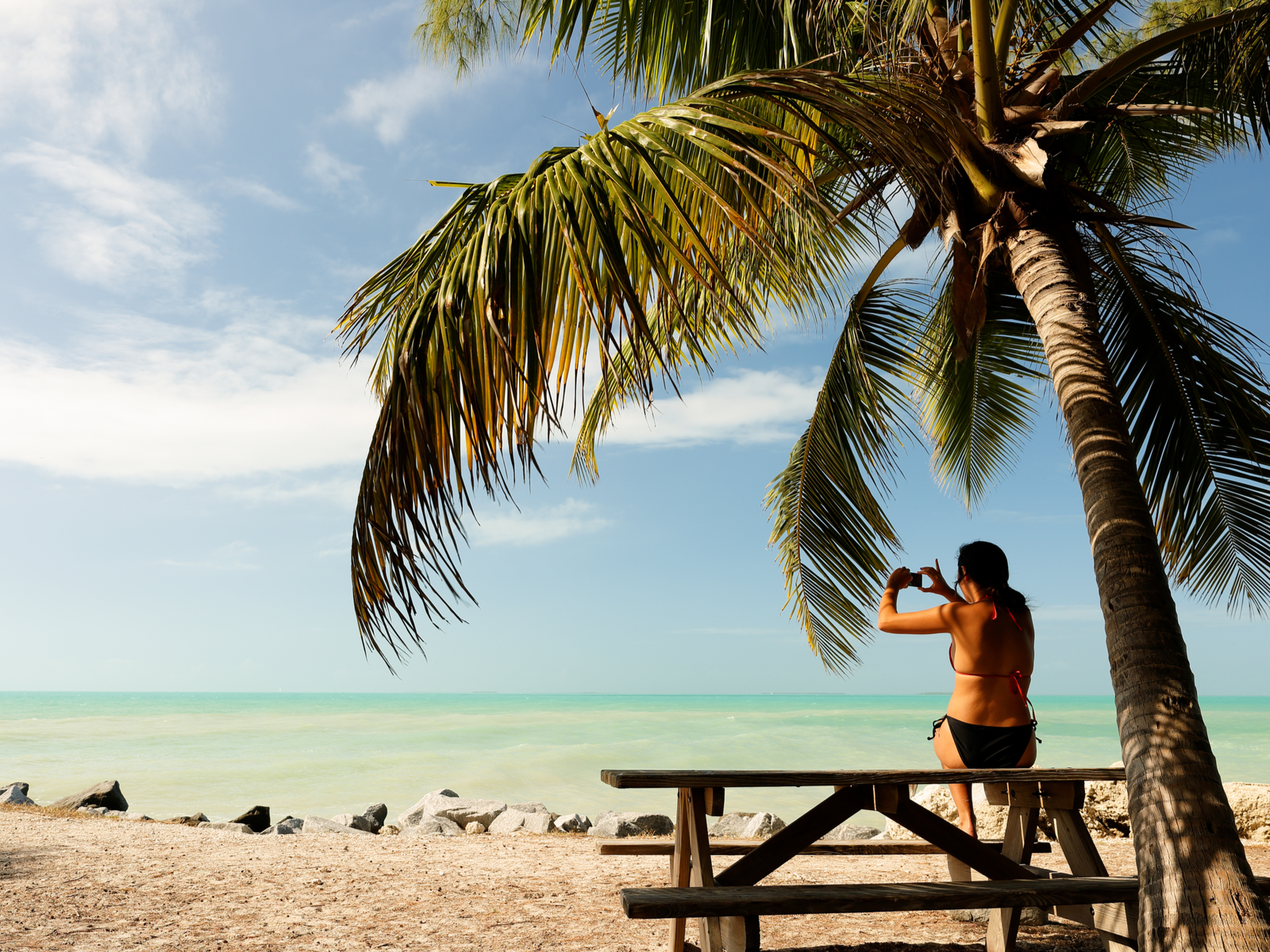 A woman wearing her swimwear sitting on a bench under a palm tree, taking a photo of the calm beach at Zachary Taylor State Park, one of the best things to do in Key West