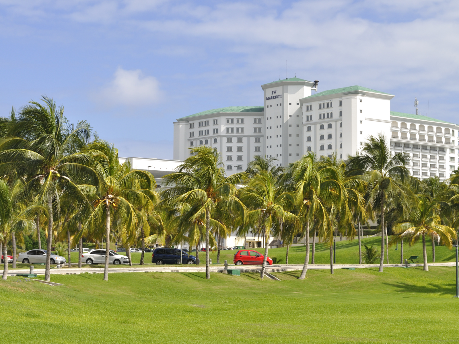 Cars driving by a small road alongside palm trees grown on green lawn with JW Marriott luxury resort and spa peaking in background, one of the best all-inclusive resorts in Cancun for families