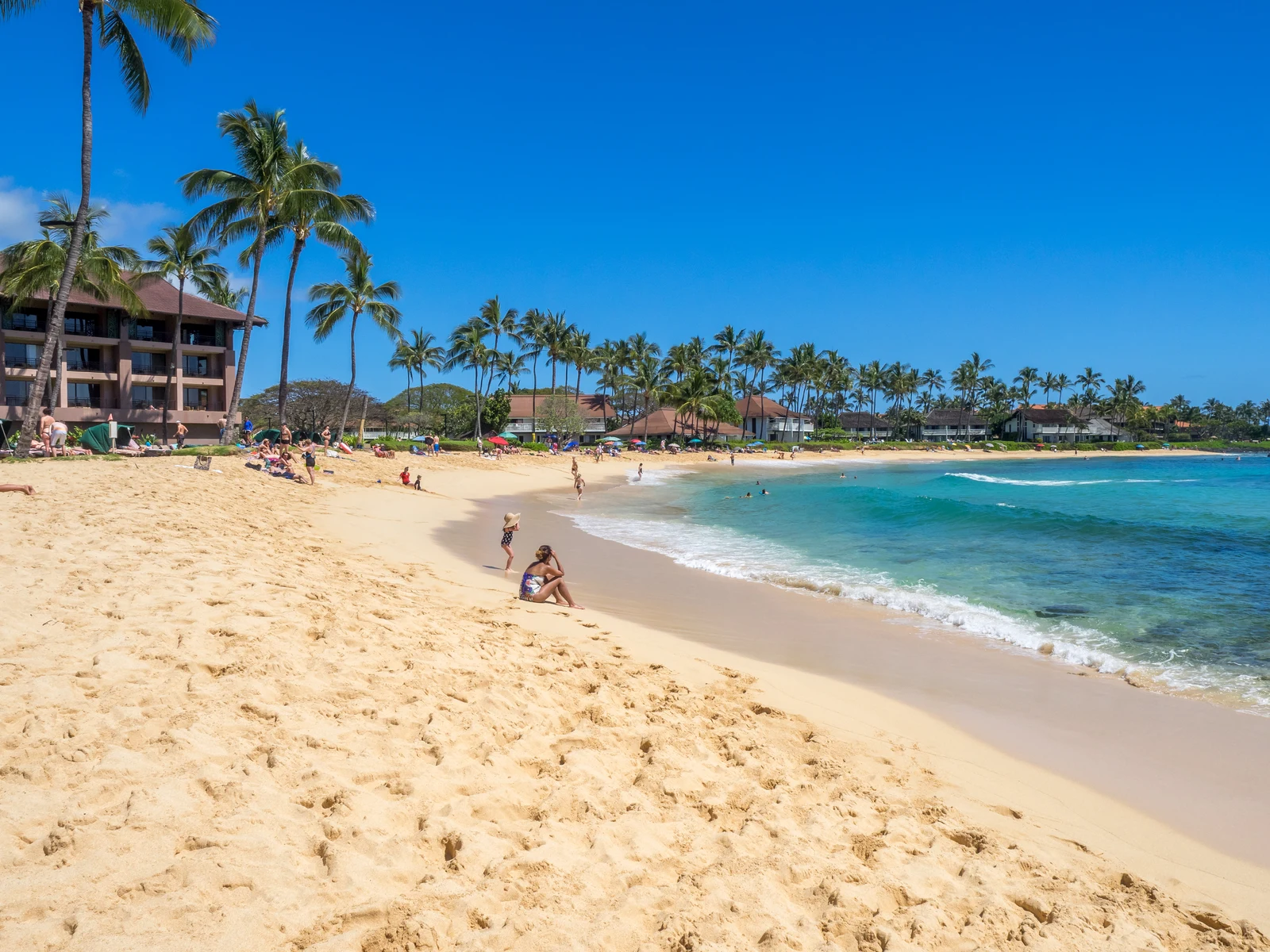Adults and kids enjoying a hot summer day at Poipu Beach, one of the best beaches in Kauai, with a series of palm trees beside hotels and resorts